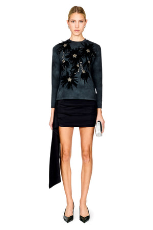 A model stands against a white background wearing a dark charcoal long-sleeve top with black floral embroidery and a black mini skirt paired with black pointed heels and holding a small silver clutch.