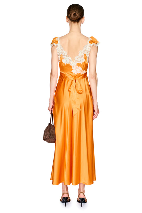 A woman shown from the back wearing a bright orange satin maxi dress with white lace shoulder details and a tie at the waist, holding a small brown handbag against a plain white background.