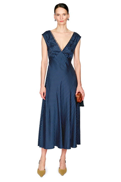 A woman modeling a sleeveless navy midi dress with a white polka dot pattern, a deep V-neckline, and cap sleeves, paired with olive green heels and holding a small brown clutch against a plain white background.