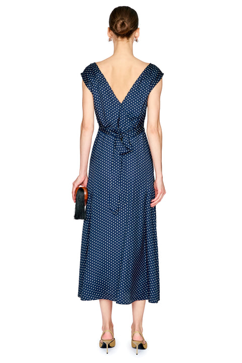 A woman viewed from the back wearing a navy midi dress with a white polka dot pattern, a wrap-style V-neckline, and a tie at the waist. She is holding a small black clutch and wearing gold heels against a plain white background.