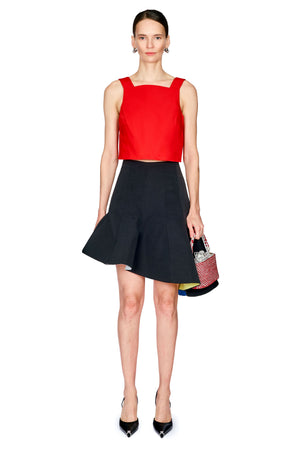 A model stands against a white background wearing a bright red sleeveless crop top and a black flared mini skirt, holding a small patterned handbag.