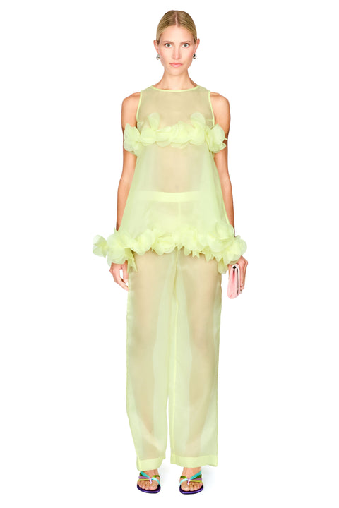 A model wearing a sheer pale yellow-green two-piece outfit with ruffled floral details on the top and wide-leg pants, paired with colorful sandals and holding a pink clutch, against a plain white background.