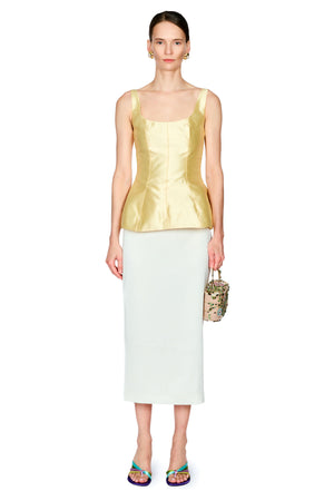 A model stands against a white studio background wearing a shiny pale yellow sleeveless top with a structured corset-style bodice and a white midi skirt, holding a small patterned handbag.