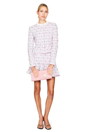 Model stands against a white background wearing a light pink and white tweed jacket and skirt set, holding a pink clutch bag