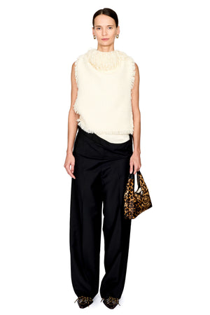 A model stands against a white studio background wearing a sleeveless cream faux-fur vest over a white top, black draped wide-leg pants, and leopard-print pointed shoes, while holding a leopard-print handbag.
