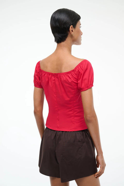 Back view of a woman wearing a bright red top with puff short sleeves that can be worn on or off the shoulder, paired with brown shorts. 