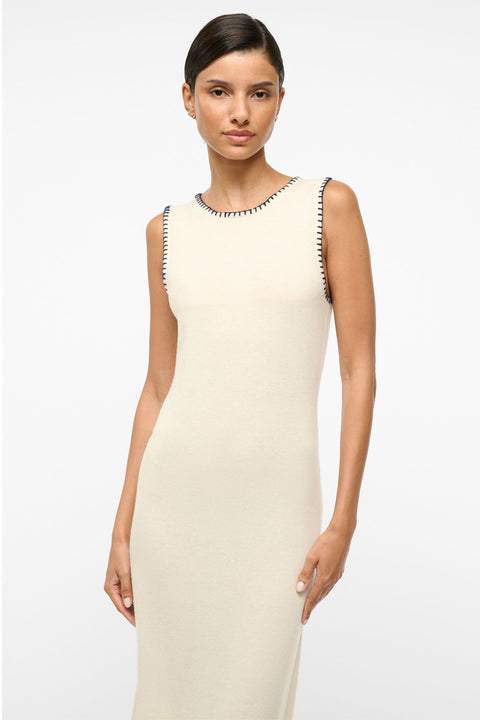 Close up of woman wearing a sleeveless ivory maxi dress that features a navy whipstitch detailing around the neck and arm holes. 