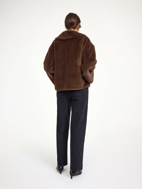 Back-facing woman wearing a brown faux fur coat that features a pointed collar, neck tie, and front pockets. 