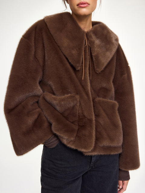 Close up of a woman wearing a brown faux fur coat that features a pointed collar, neck tie, and front pockets. 