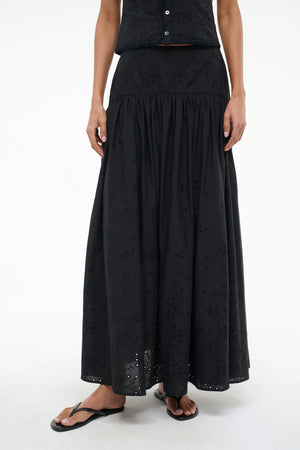 Close up of a woman wearing a black eyelet maxi skirt that features a curved yoke at the hip, paired with a matching top and black sandals. 