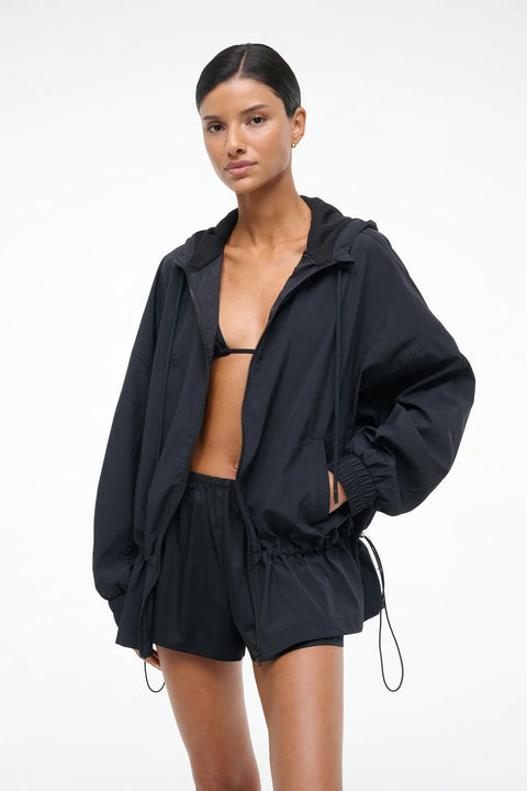 A woman wearing a pair of black poplin mini shorts with an elastic waistband, styles with a black bikini top and black windbreaker. 