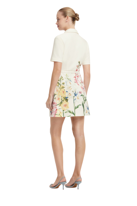 A woman shown from the back wearing a short-sleeve cream collared top and a matching cream mini skirt with a colorful floral print, paired with gray high-heeled sandals, against a plain white background.