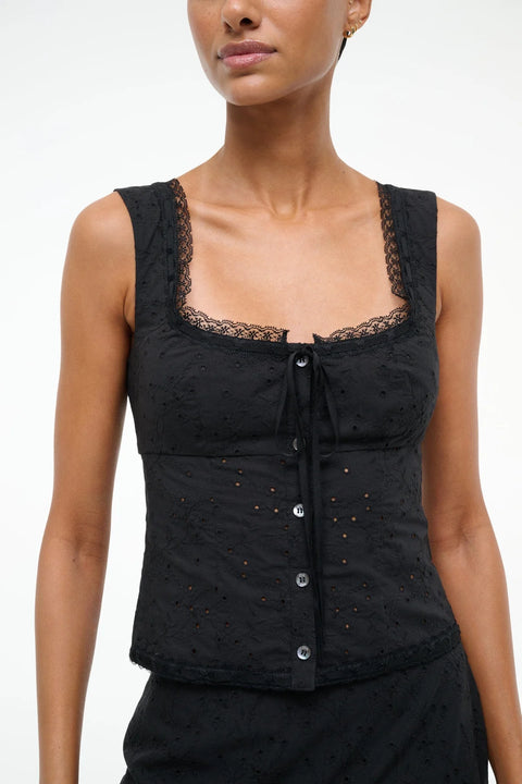 Detail shot of a woman wearing a black eyelet tank top that features lace trim, a ribbon tie neckline, and button down closure, paired with a matching skirt. 