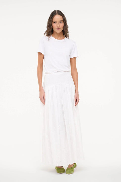 Woman wearing a white eyelet maxi skirt that features a curved yoke waist, paired with a white tee and green flats. 