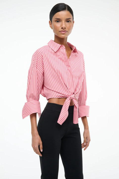 Side view of a woman wearing a cropped pink with red pinstripes button down that features cuffed sleeves and a tie at the waist, paired with black pants. 