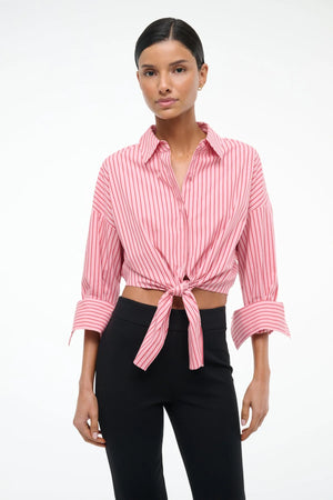 Woman wearing a cropped pink with red pinstripes button down that features cuffed sleeves and a tie at the waist, paired with black pants. 