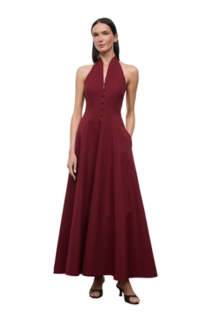 A woman modeling a sleeveless, halter-neck, floor-length maxi dress in deep burgundy. The dress features a buttoned bodice and a flowing skirt, styled with black heeled sandals against a plain white background.