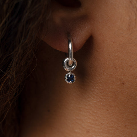 Close up of a woman wearing a sterling silver mini hoop earring that features three sterling silver annulets, one with a round blue sapphire charm. 