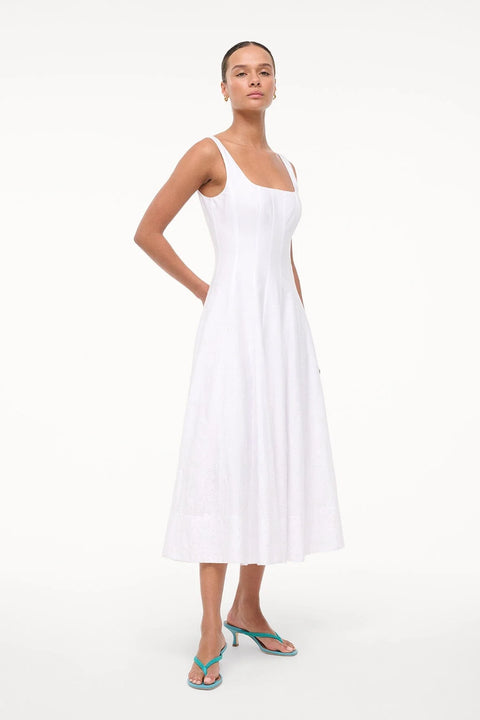 Woman wearing a sleeveless, white midi dress that features a square neck, corset style paneling, and eyelet embroidery on the A-line skirt, paired with green sandals, 