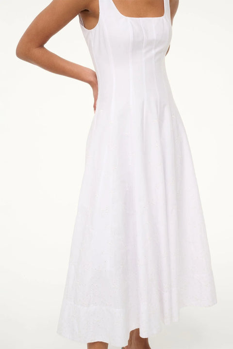Close up of a woman wearing a sleeveless, white midi dress that features a square neck, corset style paneling, and eyelet embroidery on the A-line skirt, paired with green sandals, 