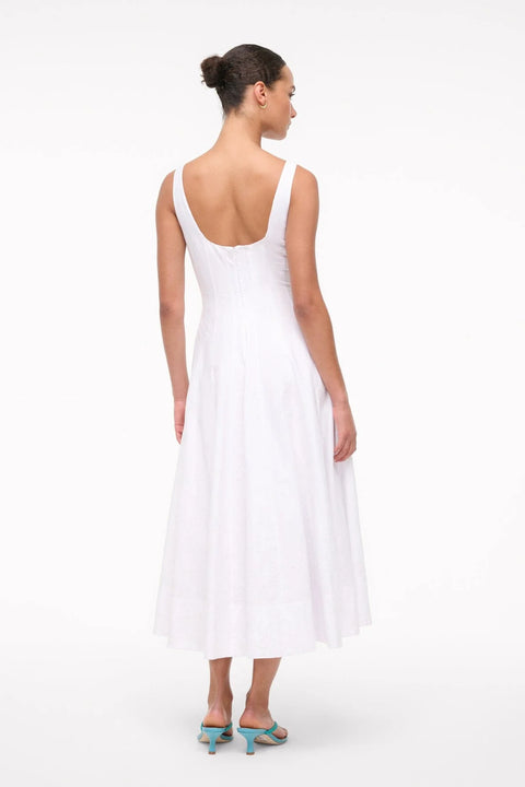 Back view of a woman wearing a sleeveless, white midi dress that features a scoop back, corset style paneling, and eyelet embroidery on the A-line skirt, paired with blue sandals.