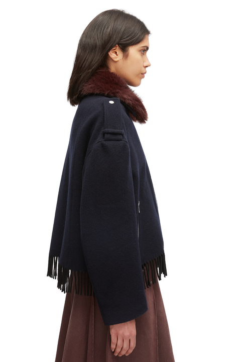A side-profile view of a model wearing a dark navy blue cape jacket with a dark brown fur collar and dark brown fringe, paired with a brown skirt, against a plain white background.