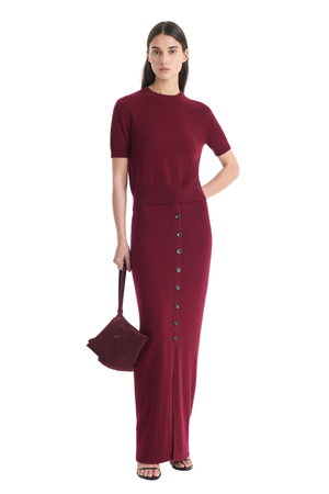 A woman modeling a long-sleeve, high-neck, floor-length knit two piece set in deep burgundy with a vertical row of dark buttons down the front. She is holding a matching burgundy handbag against a plain white background.