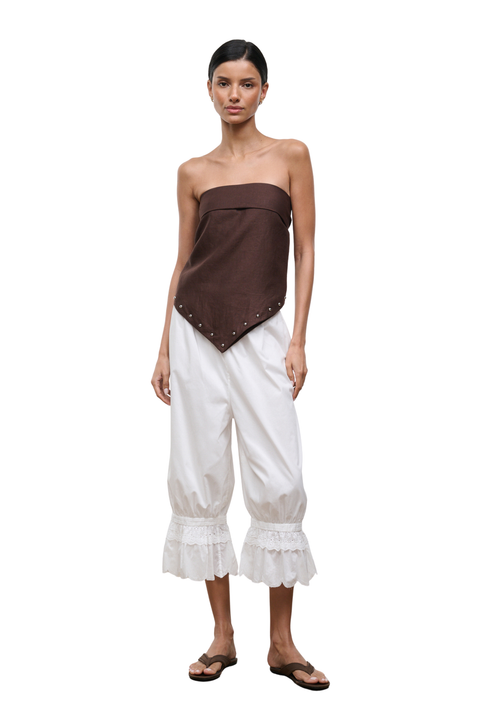 A model wearing a dark brown strapless top with silver studs and white balloon cropped pants with ruffled hems, paired with brown sandals, against a plain white background.