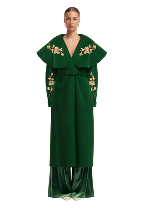 A model wearing a dark green long coat with wide lapels, gold floral embroidery on the shoulders and sleeves, a matching belt, and wide-leg trousers, shot against a solid white background.