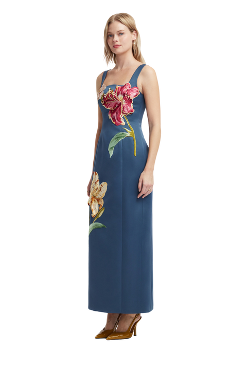 A woman modeling a sleeveless blue maxi dress with large embroidered floral details in pink, gold, and green, paired with brown pointed-toe heels against a plain white background.