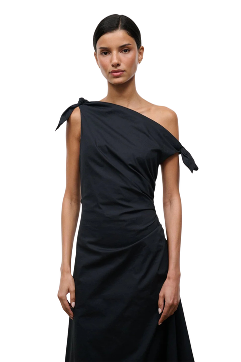 A model wearing a black one-shoulder draped dress with a tie detail on the shoulder, photographed against a solid white background.