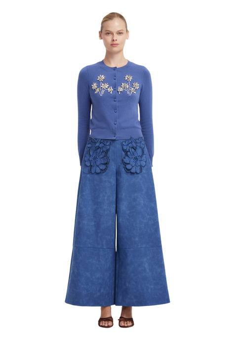 A model stands against a white background wearing a blue long-sleeve button-front top with floral embroidery on the chest and matching wide-leg pants with large floral appliqué details at the hips.