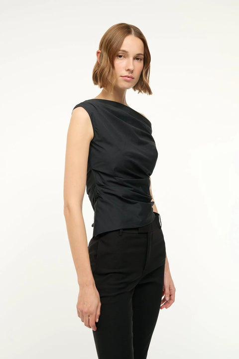 Side view of a woman wearing a black off-the-shoulder top that features asymmetrical ruching and hem, paired with black pants. 