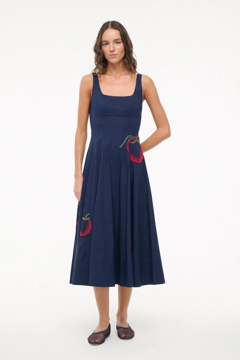 Woman wearing a sleeveless, navy midi dress that features a square neck, corset-style paneling, an A-line skirt, and red and green tomato embroidery. 