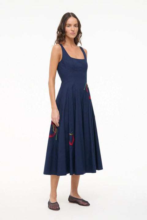 Side angle of a woman wearing a sleeveless, navy midi dress that features a square neck, corset-style paneling, an A-line skirt, and red and green tomato embroidery. 