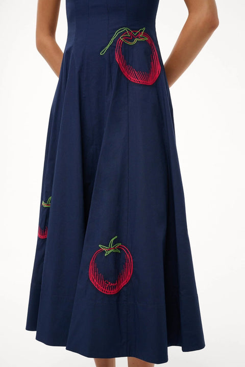 Close up of a oman wearing a sleeveless, navy midi dress that features corset-style paneling, an A-line skirt, and red and green tomato embroidery. 