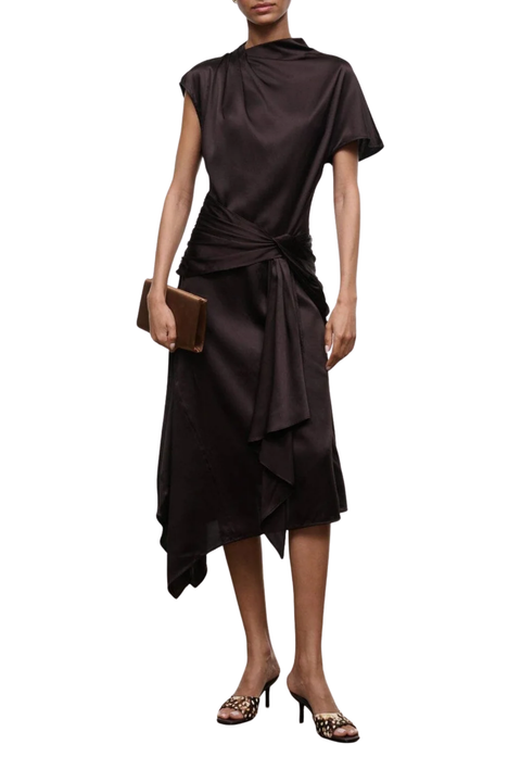 A model wearing a dark brown draped midi dress with short sleeves, a high draped neckline, and an asymmetrical hem. She is holding a small brown clutch and is against a plain white background.