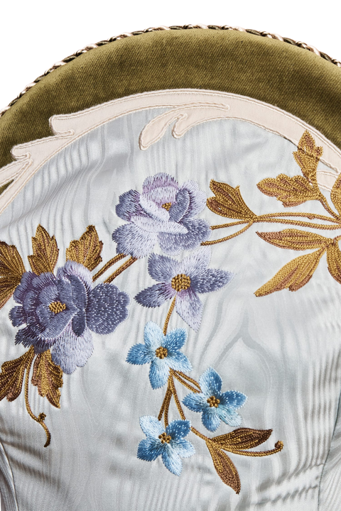 Close-up of an upholstered dress featuring a blue satin fabric with embroidered purple, blue, and gold floral motifs, topped with an olive green velvet trim.