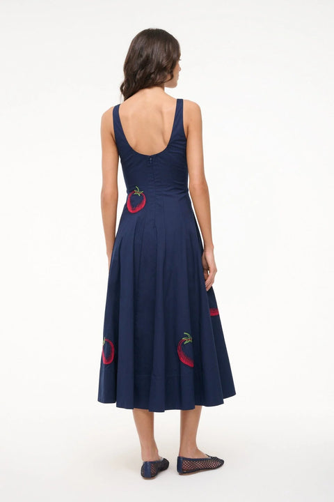 Back view of a woman wearing a sleeveless, navy midi dress that features a scoop back, corset-style paneling, an A-line skirt, and red and green tomato embroidery. 