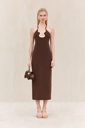 Woman wearing a brown knit midi dress that features a halter neck and an abstract Deep V accented by gold-toned hardware, paired with a brown handbag and gold sandals. 