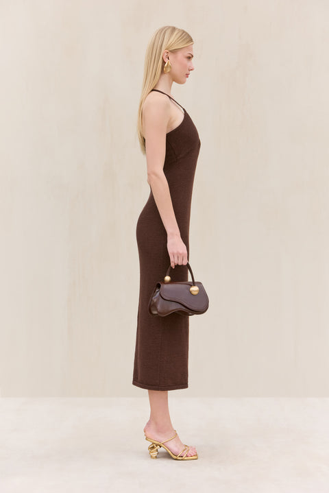 Side-facing woman wearing a brown knit midi dress that features a halter neck and an abstract Deep V accented by gold-toned hardware, paired with a brown handbag and gold sandals. 