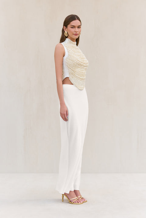 Side-facing woman wearing a cropped white tank top that features rows of pearl-beaded strands draped across the bodice, paired with a white maxi skirt. 
