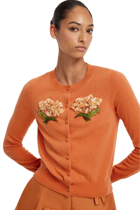 A woman wearing a burnt orange long-sleeve cardigan with embroidered floral details on the chest, paired with matching orange trousers, against a plain white background.