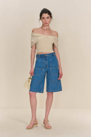 Woman wearing a tan knit off the shoulder crop top paired with blue denim shorts. 