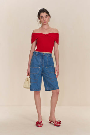 Woman wearing a red knit. off-the-shoulder, crop top paired with blue denim shorts. 