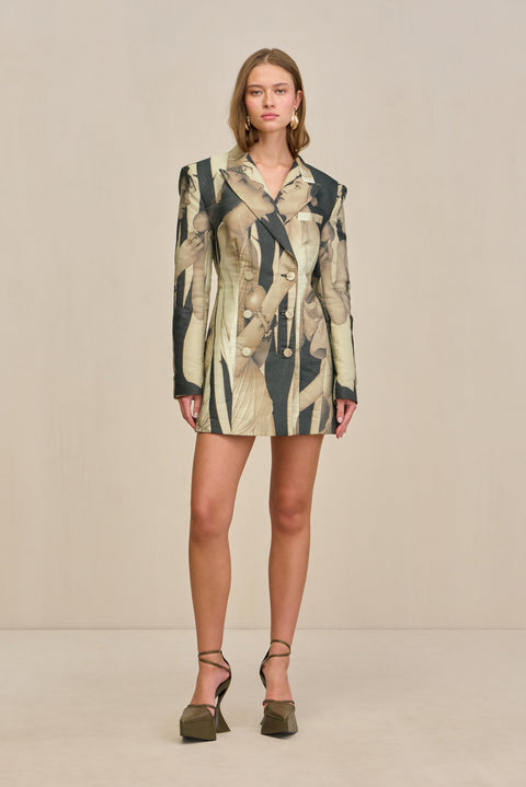 Woman wearing an oversized double-breasted blazer dress that features a black and tan portrait print and fabric covered buttons, paired with chunky oli8ve green heels. 
