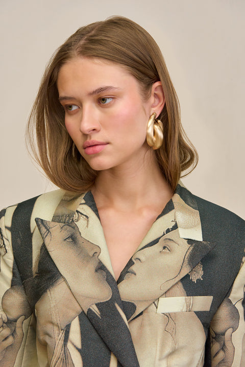 Detail shot of a woman wearing an oversized double-breasted blazer that features a black and tan portrait print depicting two women looking at each other and holding an apple, styled with chunky gold earrings. 
