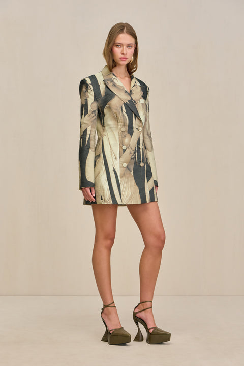 Side view of a woman wearing an oversized double-breasted blazer dress that features a portrait pattern and fabric covered buttons, styles with chunky gold earrings and bold olive green heels. 