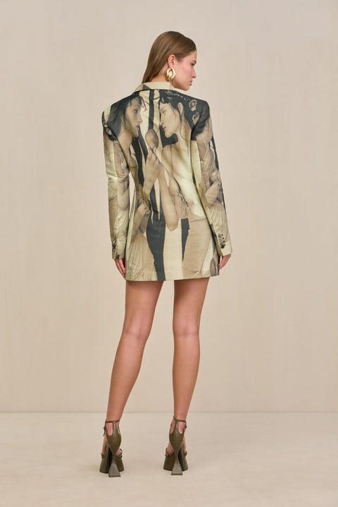 Back view of a woman wearing an oversized blazer dress that features a black and tan portrait print, paired with chunky gold earrings and olive green platform heels. 