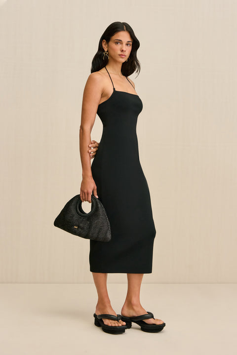 Side-facing woman wearing a black midi dress with spaghetti straps and a square neck.